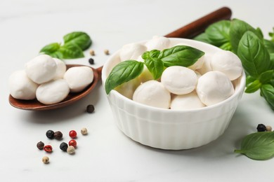 Photo of Tasty mozzarella cheese balls in bowl, spoon, basil and peppercorns on white marble table, closeup