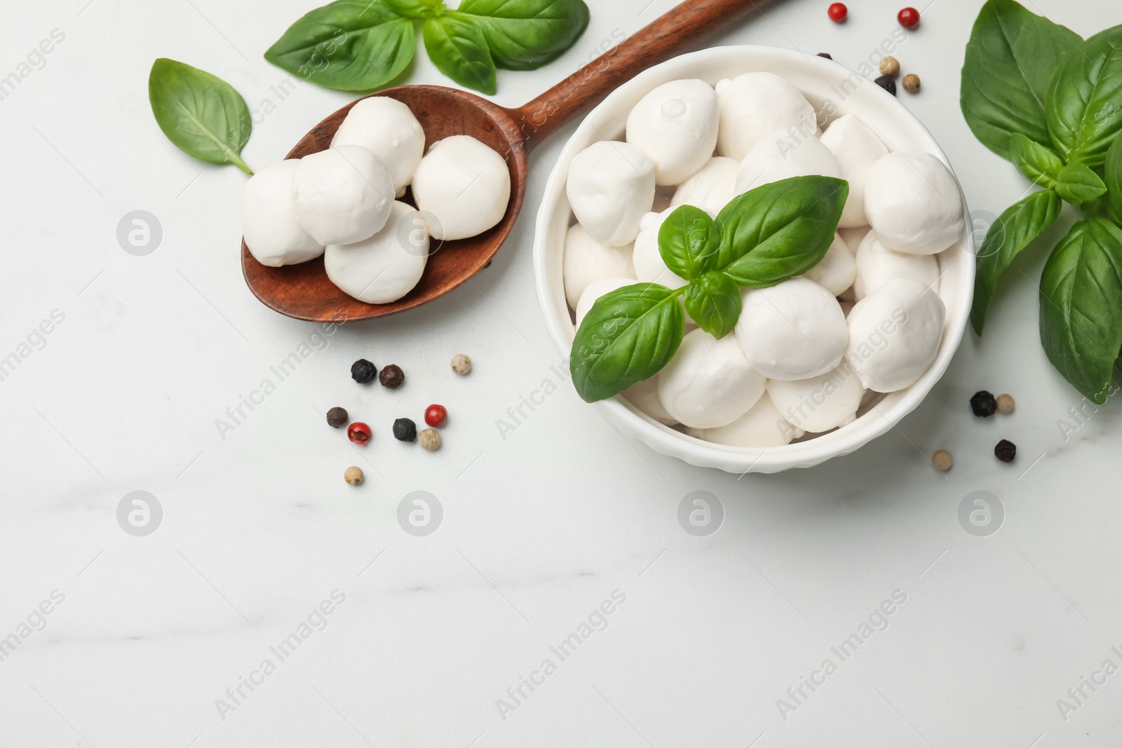 Tasty mozzarella cheese balls in bowl, spoon, basil and peppercorns on white marble table, flat lay. Space for text Photo of Tasty mozzarella cheese balls in bowl, spoon, basil and peppercorns on white marble table, flat lay. Space for text