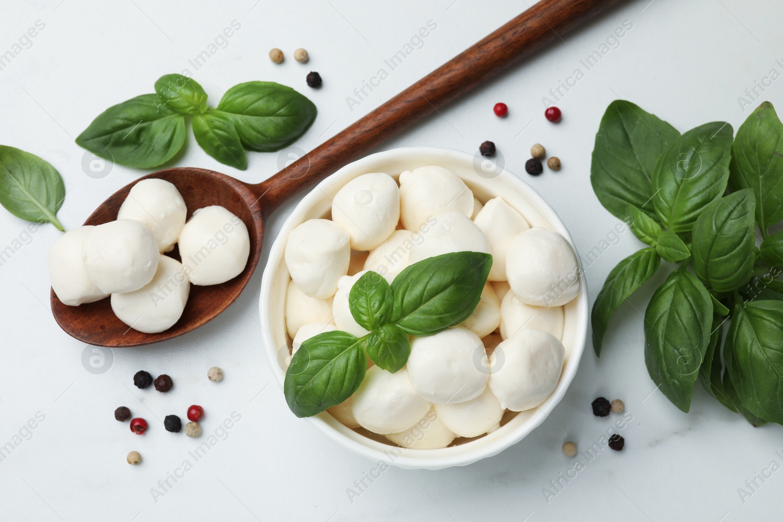Tasty mozzarella cheese balls in bowl, spoon, basil and peppercorns on white marble table, flat lay Photo of Tasty mozzarella cheese balls in bowl, spoon, basil and peppercorns on white marble table, flat lay