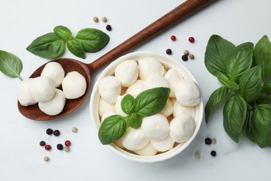 Photo of Tasty mozzarella cheese balls in bowl, spoon, basil and peppercorns on white marble table, flat lay