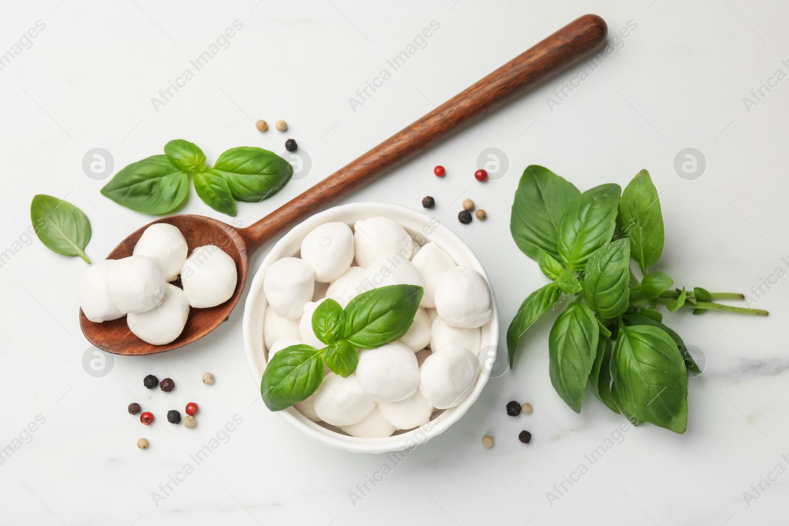 Tasty mozzarella cheese balls in bowl, spoon, basil and peppercorns on white marble table, flat lay Photo of Tasty mozzarella cheese balls in bowl, spoon, basil and peppercorns on white marble table, flat lay
