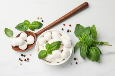 Photo of Tasty mozzarella cheese balls in bowl, spoon, basil and peppercorns on white marble table, flat lay