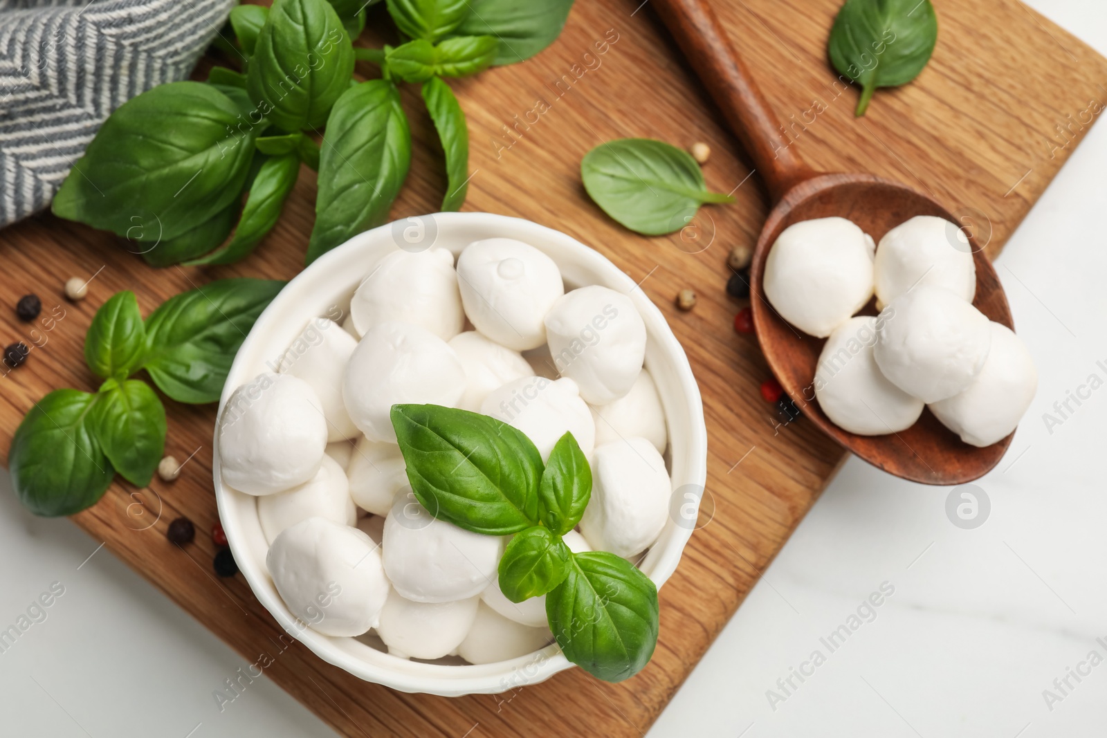 Tasty mozzarella cheese balls in bowl, spoon, basil and peppercorns on white marble table, flat lay Photo of Tasty mozzarella cheese balls in bowl, spoon, basil and peppercorns on white marble table, flat lay