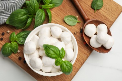 Photo of Tasty mozzarella cheese balls in bowl, spoon, basil and peppercorns on white marble table, flat lay