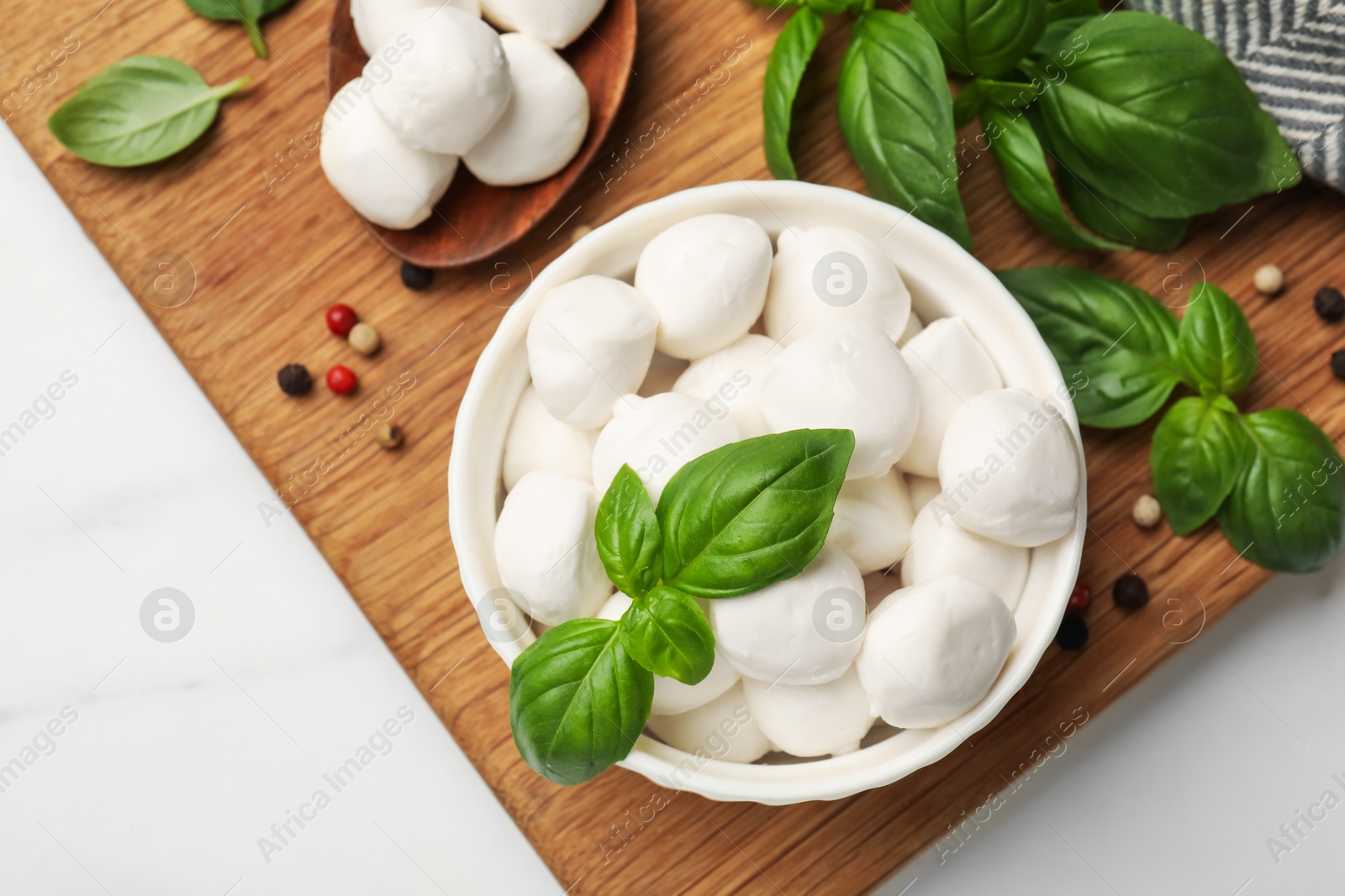 Tasty mozzarella cheese balls in bowl, spoon, basil and peppercorns on white marble table, flat lay Photo of Tasty mozzarella cheese balls in bowl, spoon, basil and peppercorns on white marble table, flat lay