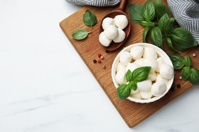 Photo of Tasty mozzarella cheese balls in bowl, spoon, basil and peppercorns on white marble table, flat lay. Space for text