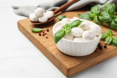 Photo of Tasty mozzarella cheese balls in bowl, spoon, basil and peppercorns on white marble table, closeup