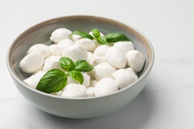 Photo of Tasty mozzarella cheese balls in bowl and basil on white marble table, closeup
