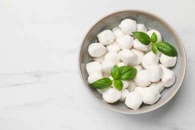 Photo of Tasty mozzarella cheese balls in bowl and basil on white marble table, top view. Space for text