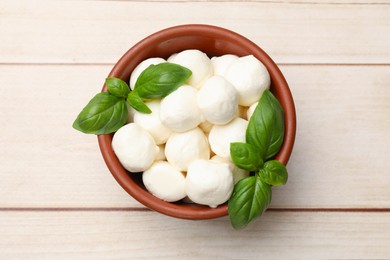 Photo of Tasty mozzarella cheese balls in bowl and basil on light wooden table, top view