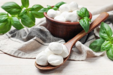 Photo of Tasty mozzarella cheese balls in bowl, basil and spoon on light wooden table, closeup