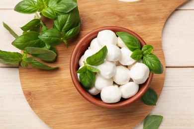 Photo of Tasty mozzarella cheese balls in bowl and basil on light wooden table, flat lay