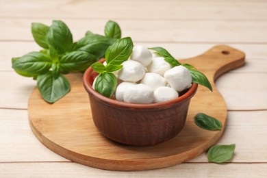 Photo of Tasty mozzarella cheese balls in bowl and basil on light wooden table, closeup