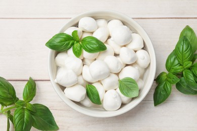Photo of Tasty mozzarella cheese balls in bowl and basil on light wooden table, flat lay