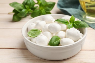 Photo of Tasty mozzarella cheese balls in bowl and basil on light wooden table, closeup
