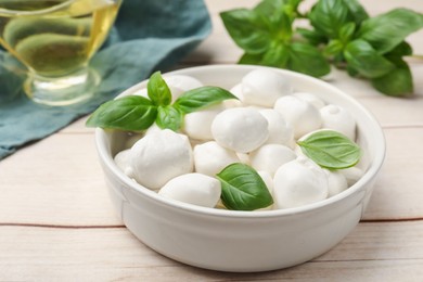 Photo of Tasty mozzarella cheese balls in bowl and basil on light wooden table, closeup