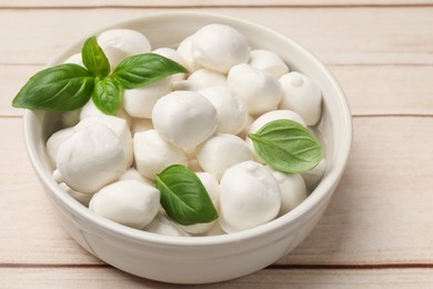 Photo of Tasty mozzarella cheese balls in bowl and basil on light wooden table, closeup