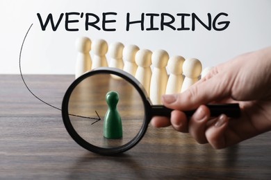 Image of We Are Hiring. Woman holding magnifying glass against green figure standing apart among wooden ones at table, closeup. Recruitment, human resources concept