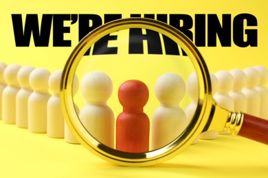 Image of We Are Hiring. Red figure among wooden ones on yellow background, view through magnifying glass. Recruitment, human resources concept