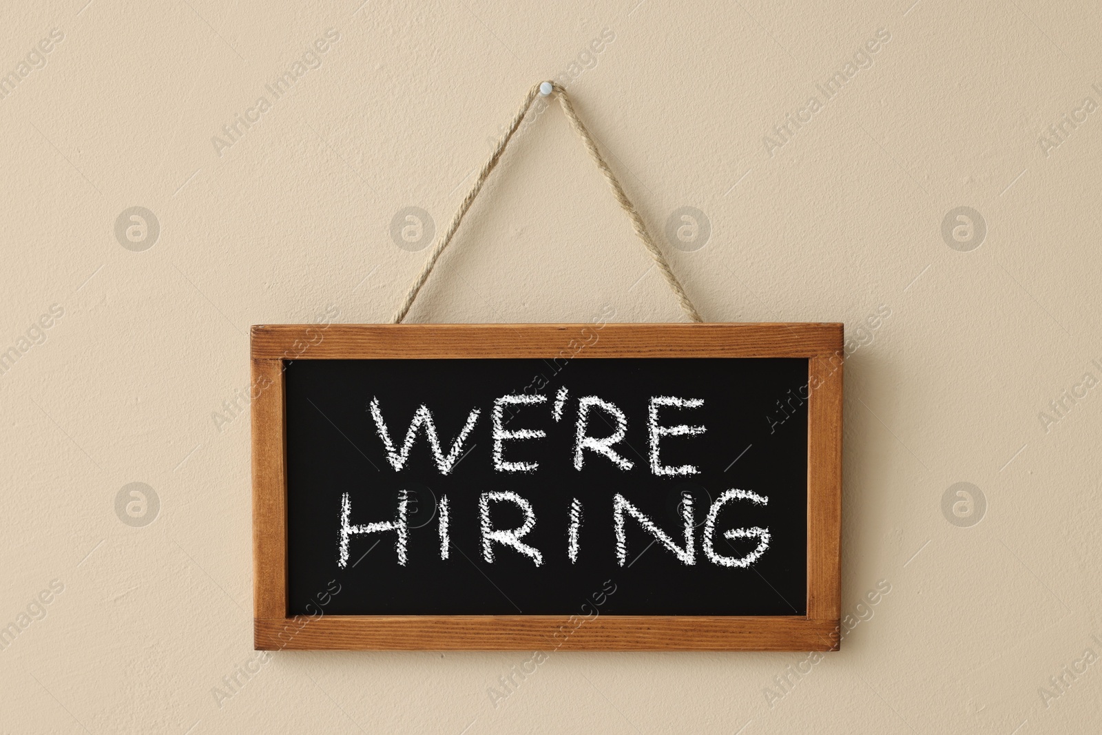 Image of Small blackboard with phrase We Are Hiring hanging on beige wall. Recruitment, human resources concept