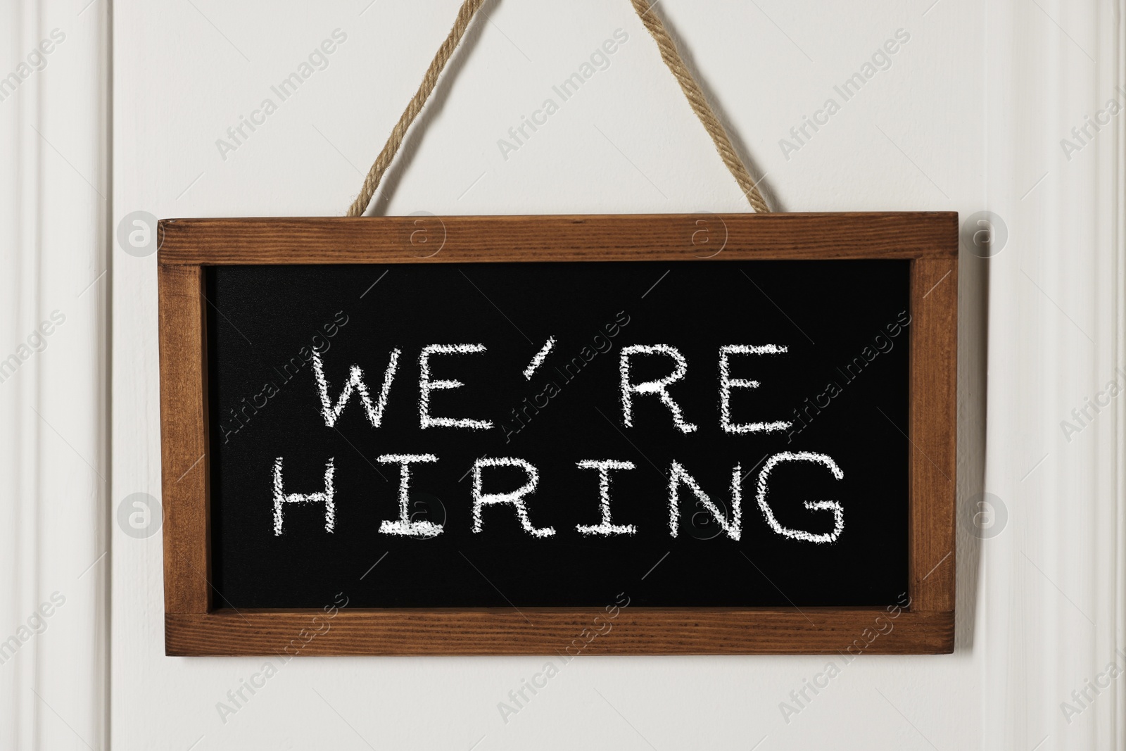 Small blackboard with phrase We Are Hiring hanging on white wall. Recruitment, human resources concept Image of Small blackboard with phrase We Are Hiring hanging on white wall. Recruitment, human resources concept