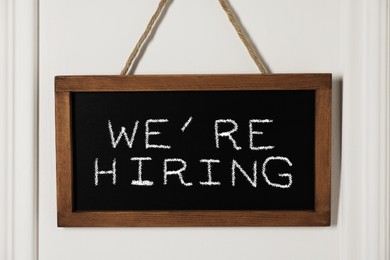 Image of Small blackboard with phrase We Are Hiring hanging on white wall. Recruitment, human resources concept