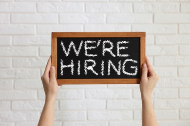 Image of Woman holding small blackboard with phrase We Are Hiring against white brick wall, closeup. Recruitment, human resources concept
