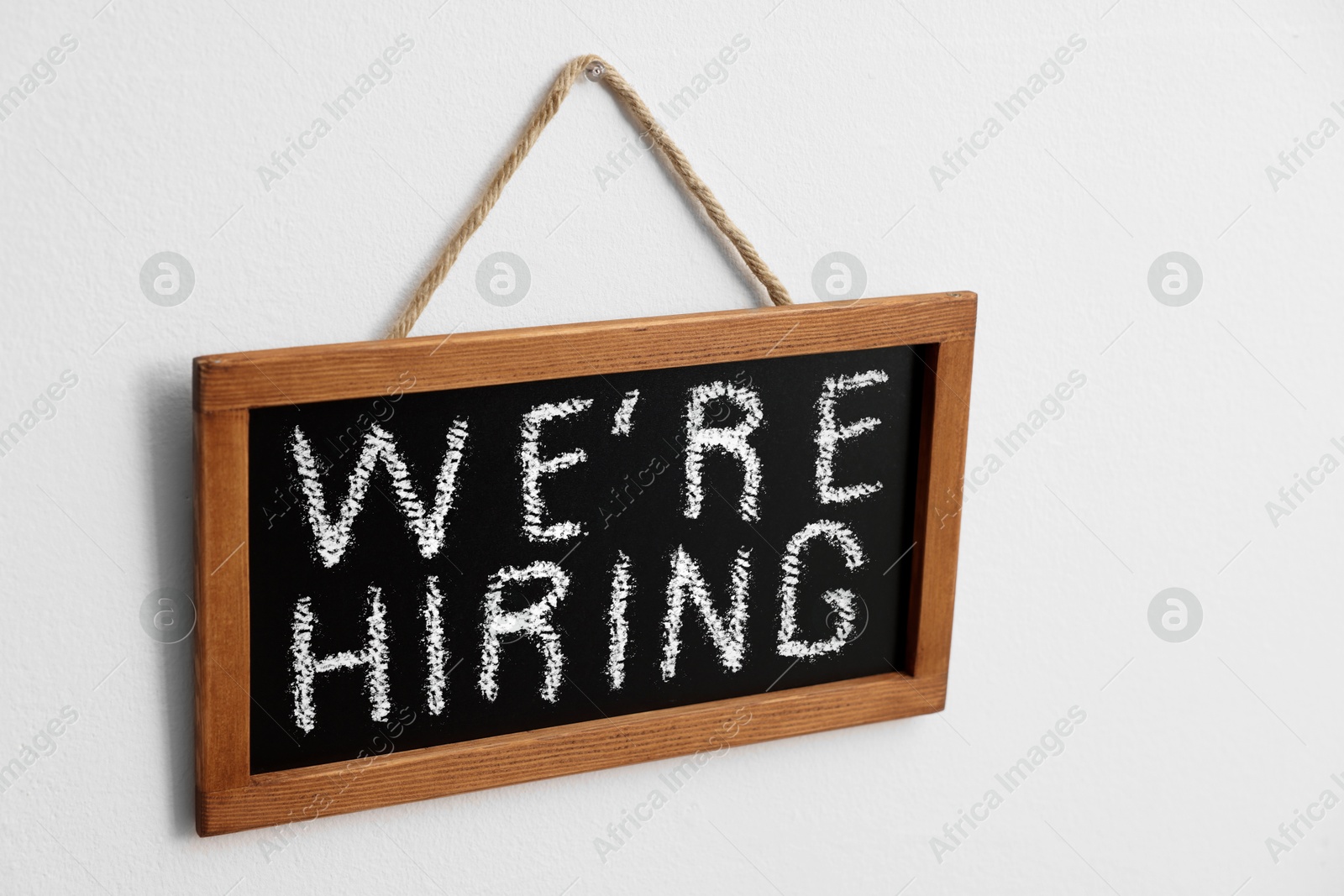 Small blackboard with phrase We Are Hiring hanging on white wall. Recruitment, human resources concept Image of Small blackboard with phrase We Are Hiring hanging on white wall. Recruitment, human resources concept
