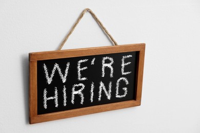 Image of Small blackboard with phrase We Are Hiring hanging on white wall. Recruitment, human resources concept