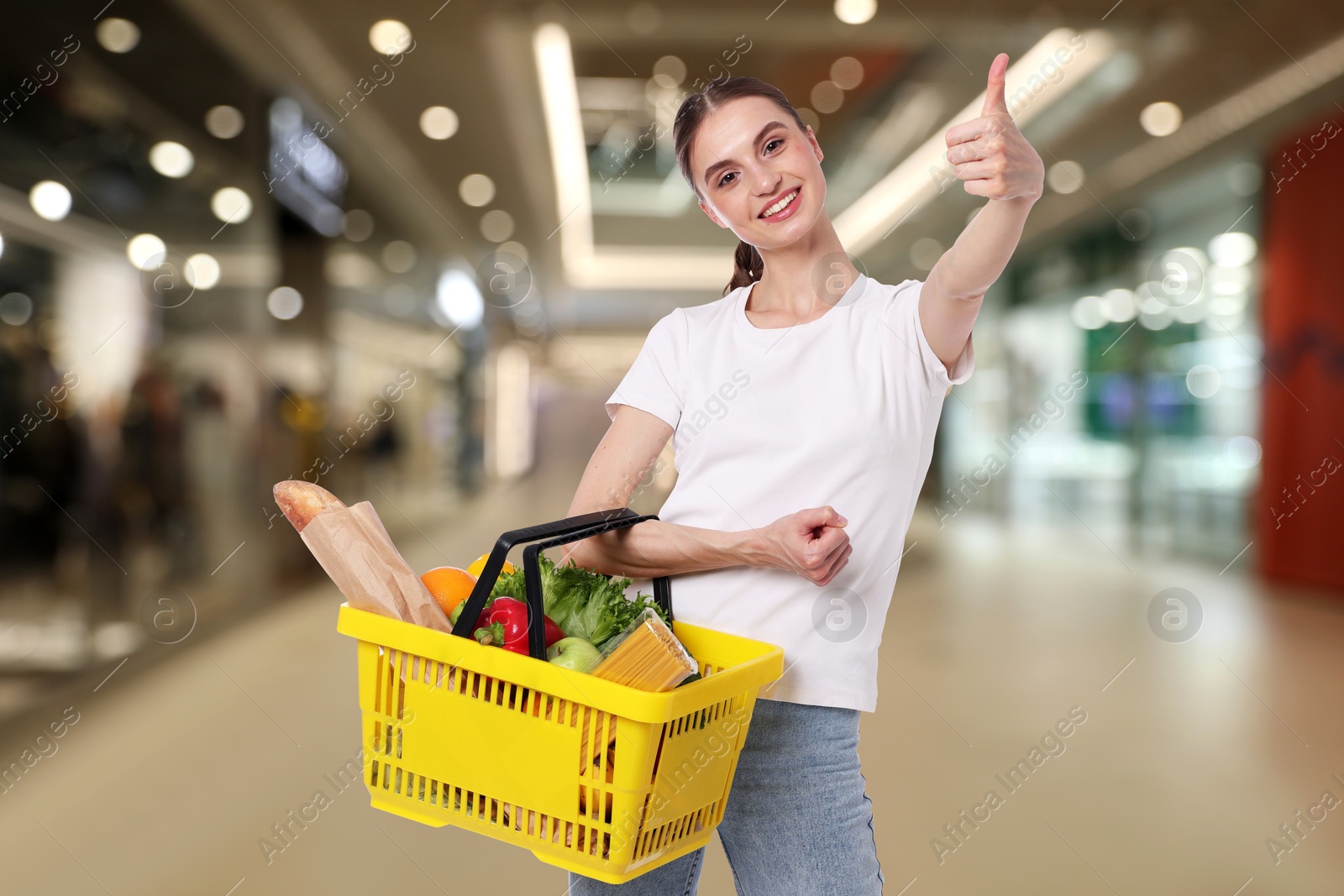 Woman holding shopping basket with different food products and showing thumbs-up at supermarket Image of Woman holding shopping basket with different food products and showing thumbs-up at supermarket