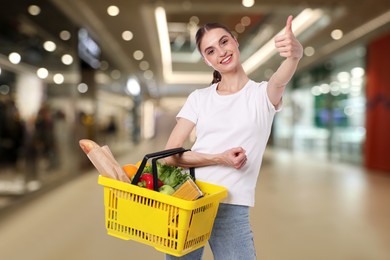 Image of Woman holding shopping basket with different food products and showing thumbs-up at supermarket