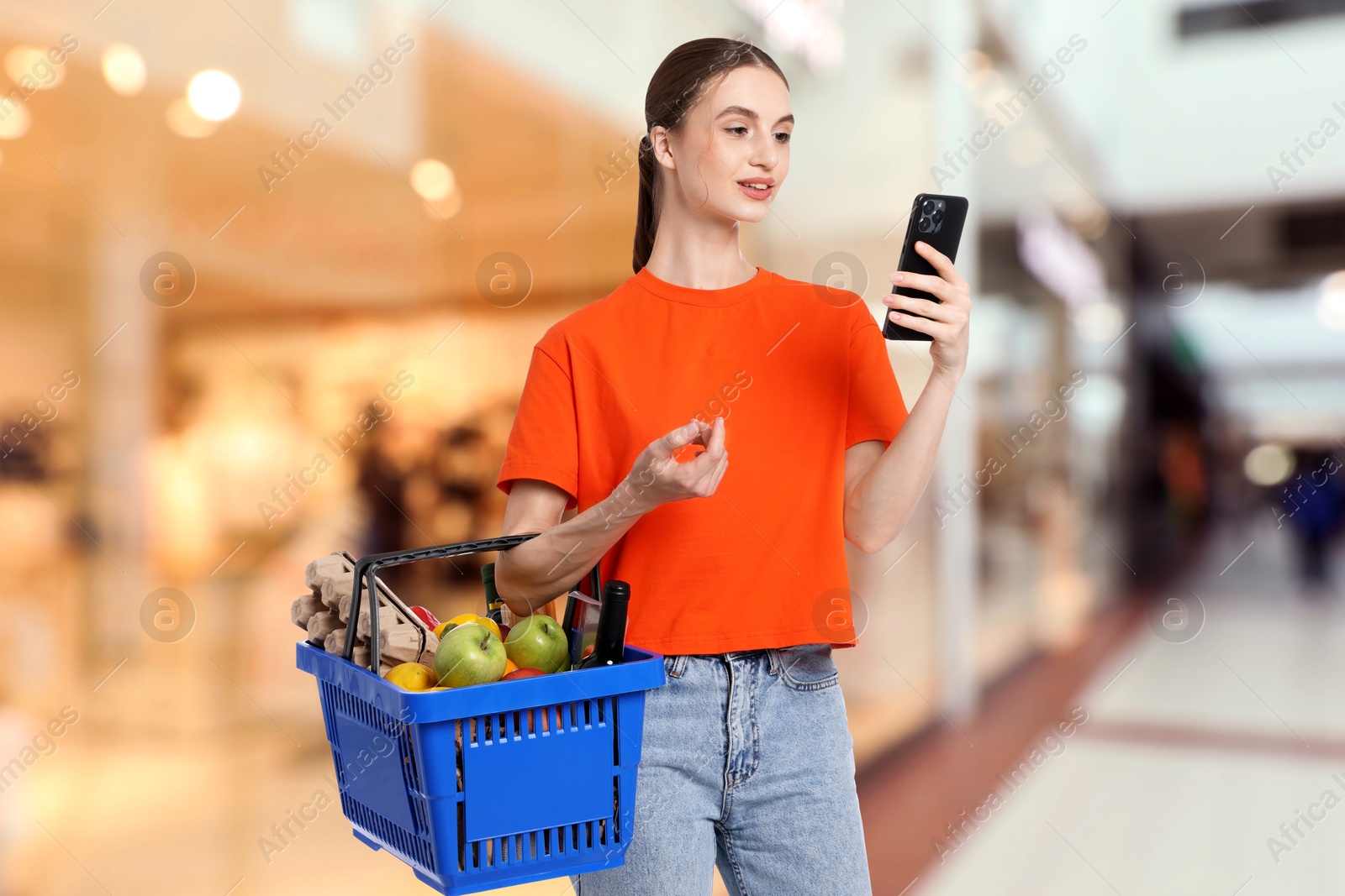 Image of Woman holding shopping basket with different food products and using mobile phone at supermarket