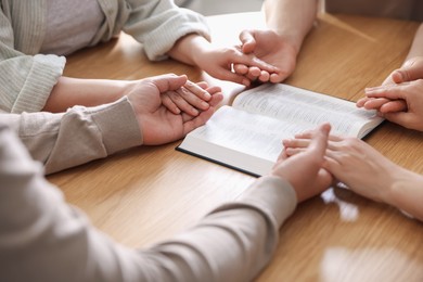 Photo of Family with Bible praying at wooden table indoors, closeup