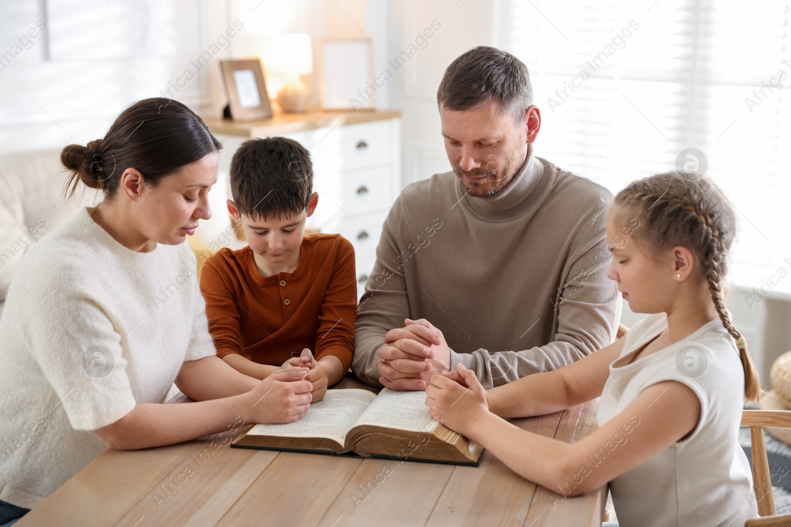 Family with Bible praying at table indoors Photo of Family with Bible praying at table indoors