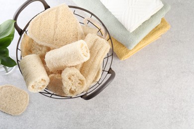 Photo of Different loofah sponges in basket and towels on light grey table, flat lay. Space for text