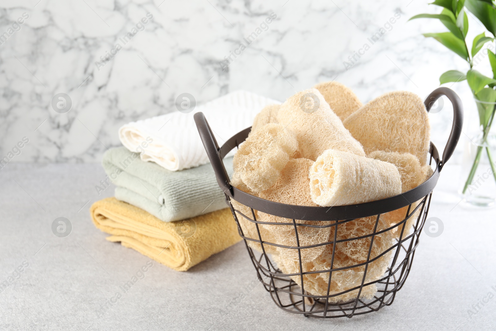 Different loofah sponges in basket and towels on light grey table Photo of Different loofah sponges in basket and towels on light grey table