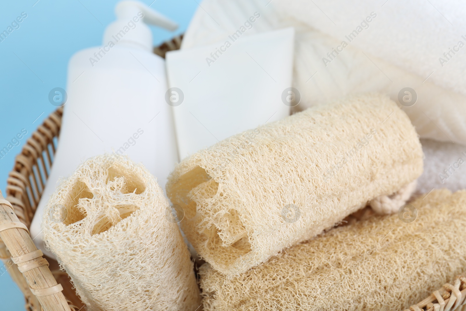 Different loofah sponges, cosmetic products and towels in wicker basket on light blue background, closeup Photo of Different loofah sponges, cosmetic products and towels in wicker basket on light blue background, closeup