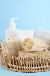 Photo of Different loofah sponges, cosmetic products and towels in wicker basket on light blue background, closeup