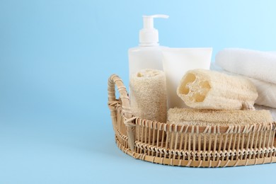 Photo of Different loofah sponges, cosmetic products and towels in wicker basket on light blue background, closeup. Space for text