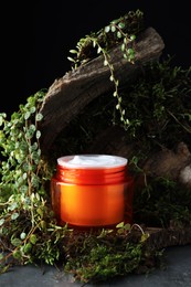 Photo of Jar of moisturizing body cream, tree bark and plants on grey table, closeup