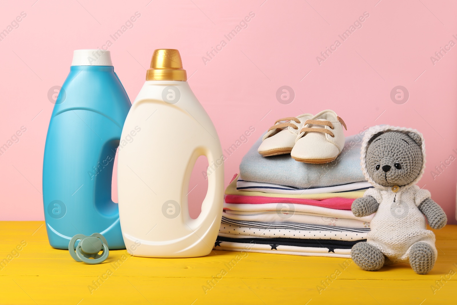 Bottles of fabric softeners, baby clothes and toy on yellow wooden table against pink background Photo of Bottles of fabric softeners, baby clothes and toy on yellow wooden table against pink background