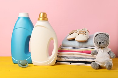 Photo of Bottles of fabric softeners, baby clothes and toy on yellow wooden table against pink background