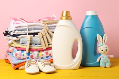 Photo of Bottles with fabric softeners, stack of baby clothes, clothespins and toy on yellow wooden table against pink background