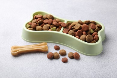 Photo of Dry pet food in feeding bowl and chew bone on gray background, closeup