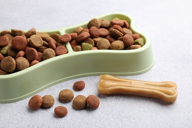 Photo of Dry pet food in feeding bowl and chew bone on gray background, closeup