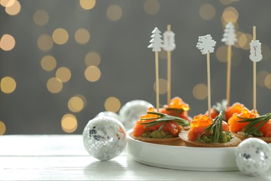 Photo of Tasty canapes and Christmas decor on white wooden table against background with blurred lights, space for text. Bokeh effect