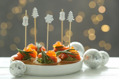 Photo of Tasty canapes and Christmas decor on white wooden table against background with blurred lights, closeup. Bokeh effect