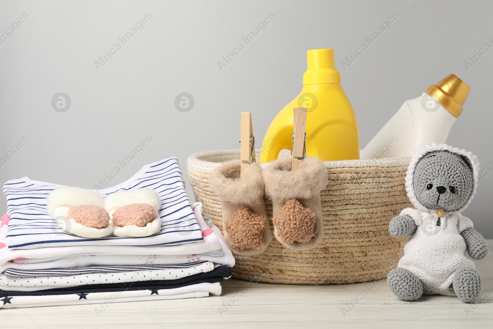 Bottles of fabric softeners, baby clothes and toy on light wooden table against grey background Photo of Bottles of fabric softeners, baby clothes and toy on light wooden table against grey background