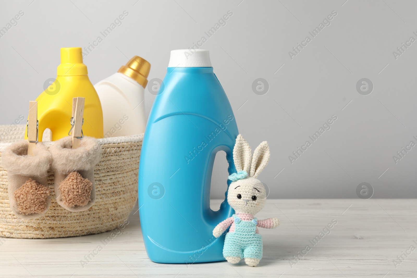 Bottles of fabric softeners, baby socks and toy on light wooden table against grey background Photo of Bottles of fabric softeners, baby socks and toy on light wooden table against grey background