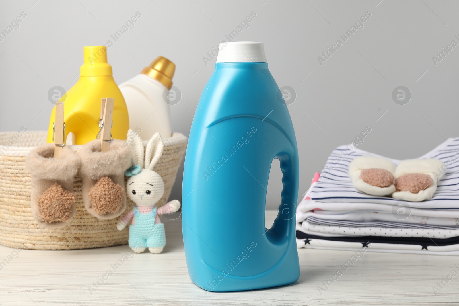 Bottles of fabric softeners, baby clothes and toy on light wooden table against grey background Photo of Bottles of fabric softeners, baby clothes and toy on light wooden table against grey background